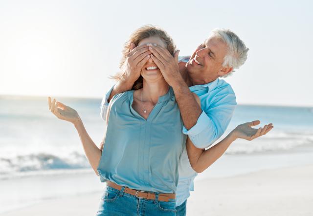 Shot of a man covering his wife's eyes while on the beach