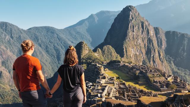 Couple holding hands standing on top of a hill, looking at the Macchu Picchu in front of them, in broad daylight.