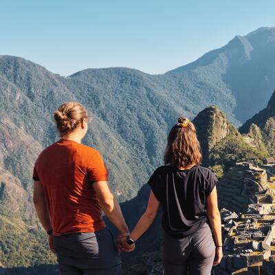 Couple holding hands standing on top of a hill, looking at the Macchu Picchu in front of them, in broad daylight.