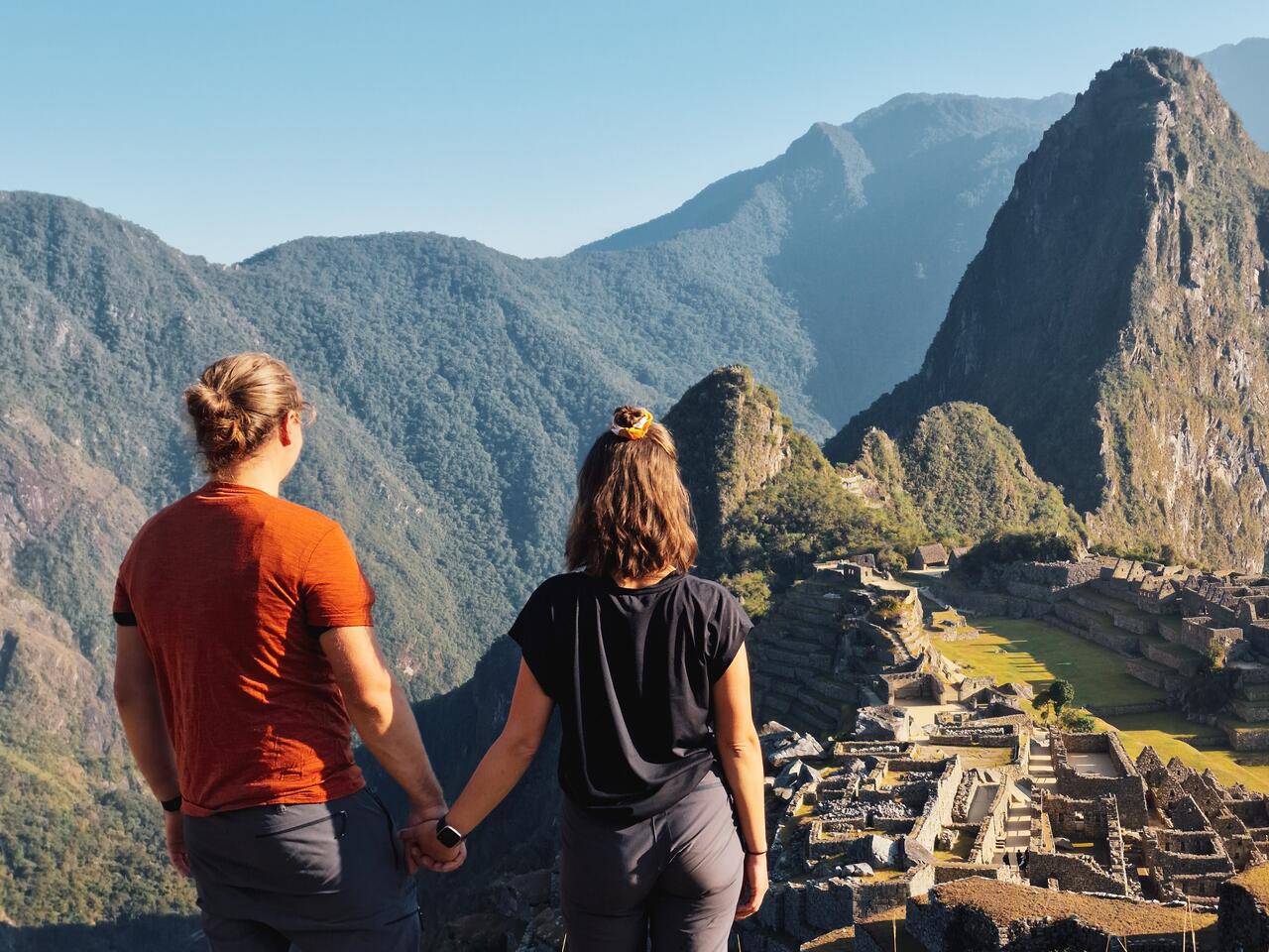 Couple holding hands standing on top of a hill, looking at the Macchu Picchu in front of them, in broad daylight.